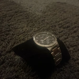 Silverfärgad klocka från Regal - Stilren klocka från Regal med svart urtavla och silverfärgat armband i metall. Urtavlan har tydliga markeringar och flera små urtavlor för extra funktioner. Klassisk rund form och länkarmband som ger en modern vibe.