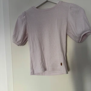 Ljuslila randig topp med puffärm Ü - Supersöt ljuslila topp från Ü med diskreta vita ränder och puffiga korta ärmar. Toppen har rund halsringning och är gjord i ett mjukt, ribbat material som känns skönt mot huden. Perfekt för dig som gillar en söt och trendig stil.