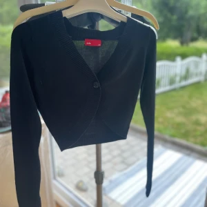 Svart gripas kofta  - Svart croppad kofta från edc. Koftan har lång ärm, ribbade muddar och en knapp framtill. Enkel och stilren design med diskret broderad logga på bröstet. 