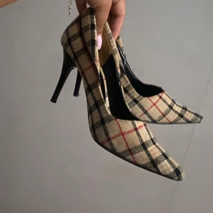 Burberry inspirerade klackskor - Snygga pumps med spetsig tå och hög klack i klassiskt rutig mönster. Skorna har beige bas med svarta och röda rutor, samt svart klack. Yttermaterialet är tyg och ger en trendig vibe till outfiten. Fint sckik inga synliga täcken på sliten användning, 