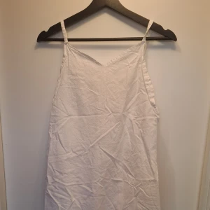 Vit somrig klänning med smala axelband - Enkel och luftig vit klänning med smala axelband och v-ringad hals. Klänningen är ärmlös och har en rak, avslappnad passform. Perfekt för varma dagar och lätt att styla med accessoarer. Materialet känns svalt och passar till sommaren. Vet inte riktigt hur många gånger den är använd men de ser ut att vara i bra skick.