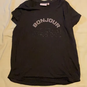 Svart t-shirt BONJOUR från R-COVER - Svart t-shirt från R-COVER med trycket 'BONJOUR' i vitt och glittrande detaljer under texten. Klassisk rund hals och korta ärmar. Perfekt för dig som gillar enkel men snygg stil.