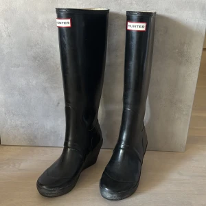Svarta Hunter stövlar med kilklack - Snygga svarta stövlar från Hunter med klassisk logga framtill. Höga skaft och kilklack ger en cool siluett. Tillverkade i slitstarkt gummi som håller dig torr i regnet. Perfekta för dig som vill ha både stil och funktion under ruggiga dagar.
