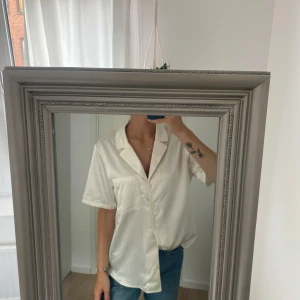 Vit kortärmad skjorta i satin-look - Snygg vit skjorta med kort ärm och avslappnad passform. Skjortan har klassisk krage, bröstficka och knäppning framtill. Materialet har en lätt glansig satin-feel som ger en fräsch vibe. Perfekt till jeans eller kjol för en clean och stilren look.