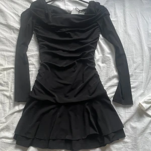 Svart kort klänning  - Supersnygg  svart kort klänning från Bershka med långa ärmar och draperad effekt över hela kroppen. Klänningen har en volangig kjol som ger extra rörelse. Perfekt för dig som vill sticka ut med en stilren look.
