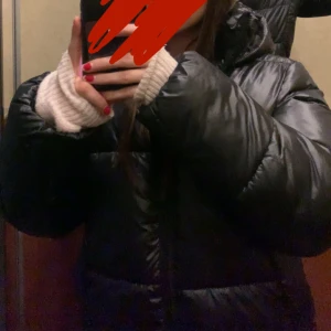 Svart pufferjacka med huva - Säljer en svart pufferjacka med huva, perfekt för kalla dagar. Jackan har en glansig finish och är riktigt fluffig och varm. Stängs med dragkedja framtill och har långa ärmar. Enkel och stilren design som passar till det mesta.