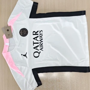 PSG x Jordan vit och rosa fotbollströja - Snygg PSG fotbollströja i vitt med svarta och rosa detaljer. Tröjan har korta ärmar, svart Jordan-logga och Paris Saint-Germain-emblem på bröstet. Materialet är lätt och andas, perfekt för träning eller match. Matchande svarta shorts med rosa inslag och loggor ingår.