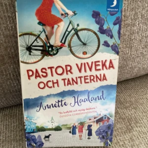 Pastor Viveka och tanterna - En charmig bok med färgstark omslagsbild där en kvinna cyklar och tanter samlas i en idyllisk bymiljö. Perfekt för dig som gillar mysiga berättelser med humor och spänning. Pocketutgåva som passar unga vuxna som vill ha lättsam och underhållande läsning.