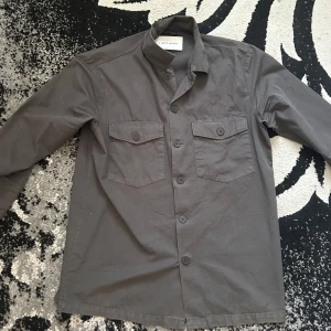 Grå overshirt från A Day's March - Säljer en stilren grå overshirt från A Day's March i storlek small. Skjortan har två bröstfickor med lock och knappar, klassisk krage och långa ärmar. Perfekt att bära öppen eller stängd för en avslappnad look.