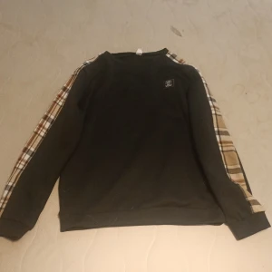 Svart sweatshirt med rutiga ärmar från burberry - Svart sweatshirt med rutiga detaljer längs ärmarna i beige, brunt och vitt. Tröjan har rund halsringning och en liten patch på bröstet. Perfekt för dig som gillar en chill och trendig stil., burberry