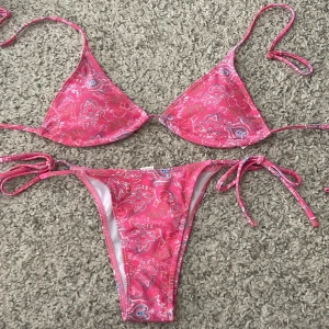 Rosa bikini med paisleymönster och knyt - Snygg rosa bikini med paisleyinspirerat blommönster i vitt, blått och rött. Klassisk trekantsmodell upptill och knyt i sidorna på underdelen. Perfekt för stranden eller poolen, med tunna band och skön passform. Inte använd. Skriv för frågor. 