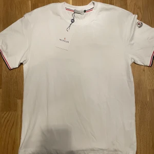 Vit Moncler t-shirt med logga - Vit t-shirt från Moncler i mjuk bomull. Klassisk passform med korta ärmar och rund hals. Ärmsluten har snygga ränder i rött och blått, och vänster ärm pryds av Monclers ikoniska logga. Perfekt för en clean och stilren look.