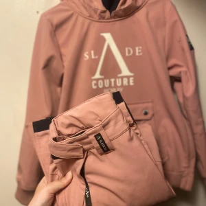 Rosa vindjacka från Slade Couture - Snygg rosa vindjacka från Slade Couture med stor vit logga framtill och huva. Jackan har praktisk ficka med knapp och är perfekt för dig som gillar streetstyle. Materialet känns mjukt och sportigt, och jackan har långa ärmar. Tillhörande byxor i samma material. Storlek 150kr. Nypris 1500kr, ligger nu på rea för 799kr för bara jackan. Säljer BÅDA för 550kr.
