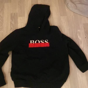 Svart hoodie från Hugo Boss med tryck - Svart hoodie från Hugo Boss med stor huva och vit BOSS-logga framtill, understruken med rött tryck. Klassisk passform med långa ärmar och mjukt material som känns skönt mot huden. Perfekt för dig som gillar stilrena streetwear-plagg.