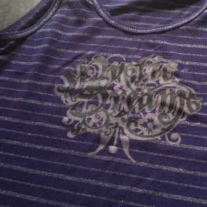 Lila randig linnetopp från mywear - Lila ärmlös linnetopp från mywear med silvriga horisontella ränder och tryck framtill i grått och svart. Toppens material är mjukt och stretchigt, perfekt för en avslappnad stil. Rund halsringning och smala axelband ger en klassisk look.