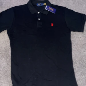 Svart pikétröja från Polo Ralph Lauren - Klassisk svart pikétröja från Polo Ralph Lauren med röd broderad logga på bröstet. Tröjan har korta ärmar, krage och knappar framtill. Tillverkad i mjuk bomull som känns skön mot huden. Perfekt för en stilren och avslappnad look.