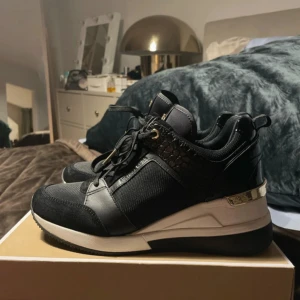 Michael Kors skor - Snygga svarta sneakers från Michael Kors med gulddetaljer och logga på plösen. Skorna har en mix av skinn, mocka och textil, samt en vit och svart sula med guldfärgad detalj vid hälen. Perfekta för dig som gillar en sportig men lyxig look.
