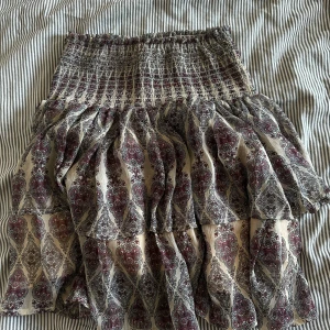 Mönstrad kortkjol från Sofie Schnoor - Supersöt kortkjol från Sofie Schnoor med volanger och smock i midjan. Kjolen har ett detaljerat paisleymönster i vinrött, svart och beige på ljus botten. Perfekt för dig som gillar boho-vibes och vill ha något luftigt och snyggt till sommaren. Aldrig använd! 🩷