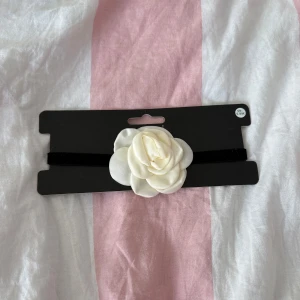 Choker med vit blomma från Ur&Penn - Supersnygg svart sammetschoker från Ur&Penn med en stor vit tygblomma framtill. Halsbandet har justerbart spänne i silverfärg och ger en cool och romantisk vibe. Perfekt accessoar för att lyfta din outfit med en unik detalj.