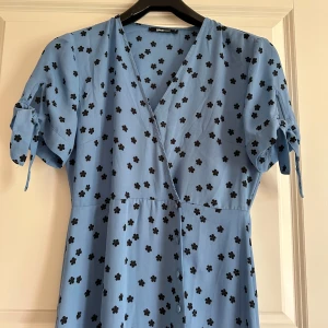 Blommig blå omlottklänning Gina Tricot - Ljusblå långklänning med svarta blommor från Gina Tricot. Klänningen har omlott-skärning, v-ringning och korta ärmar med knyt. Perfekt till sommarens alla tillfällen för en söt look. Aldrig använd.