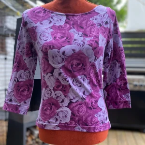Blommig topp med lila rosor - Handsydd topp med rund halsringning och 3/4 ärmar. Den har ett blommigt tryck över hela plagget. Längd: 53 cm ﻿bystvidd: 53 cm ﻿Bomull/elastan  Inga bilder när den sitter på mig! Katter finns i hemmet!