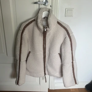 Offwhite teddyjacka från H&M XS - En Stor Favorit! Supermjuk teddyjacka från H&M i offwhite med bruna detaljer längs dragkedjan och ärmarna. Jackan har hög krage, två fickor och hel dragkedja framtill. Perfekt för kalla dagar och ger en chill vibe till din outfit. I mycket fint skick, använd  typ 2 ggr! Har en beige också så denna blev mest bara hängandes!