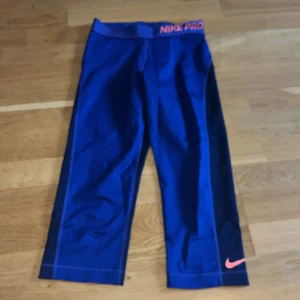 Blå Nike Pro träningstights - Säljer ett par blå Nike Pro träningstights (går över knäna)  med orange Nike-logga på benet och Nike Pro-tryck i midjan. Tightsen är trekvartsmodell och har en tight passform, perfekta för träning.