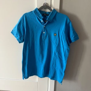 Blå pikétröja från Lyle & Scott - Säljer en blå pikétröja från Lyle & Scott med klassisk krage och knappslå. Tröjan har korta ärmar och den ikoniska gula loggan broderad på bröstet. Perfekt för en stilren och avslappnad look. Använd - i gott skick 