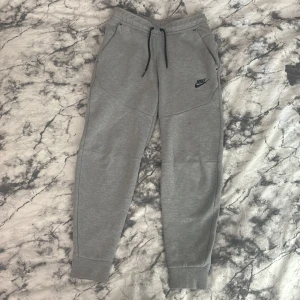Grå mjukisbyxor till nike tech fleece set - Säljer ett par grå mjukisbyxor från Nike med svarta dragsnören och logga på vänster ben. Byxorna har fickor fram och en bakficka med dragkedja. Perfekta för chill eller träning.