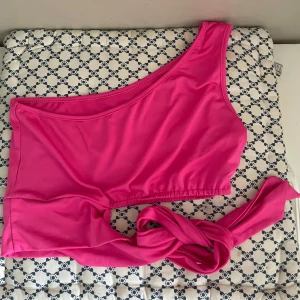 Rosa one shoulder topp med knyt - Säljer en snygg rosa one shoulder topp med knyt i sidan. Toppen är croppad och har en modern, asymmetrisk design som passar perfekt till sommaren.