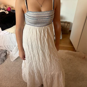 Vit och blå sommarklänning från Zara - Säljer en söt vit långklänning från Zara med smock upptill i blått och vitt. Klänningen har smala axelband och en luftig, flowy passform – perfekt för varma dagar. Tillverkad i ett lätt material som känns skönt mot huden.