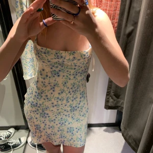 Blommig axelbandslös kortklänning - Söt axelbandslös kortklänning med blommigt mönster i blått och grönt på ljus botten. Figurnära passform och perfekt för varma dagar. Klänningen har en enkel och fräsch design som passar till många tillfällen.