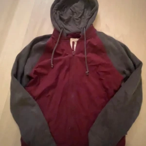 Bordeaux och grå hoodie från Hollister - Säljer en snygg hoodie från Hollister i bordeauxrött med grå ärmar och huva. Tröjan har dragsko i huvan och är perfekt för en avslappnad stil. Klassisk passform och mjukt material.