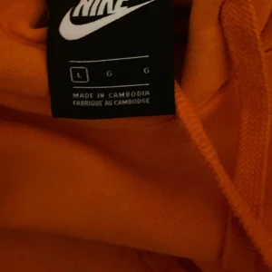 Orange hoodie från Nike - Säljer en orange hoodie från Nike i storlek L. Tröjan har huva med dragsko och en klassisk Nike-logga broderad på bröstet. Perfekt för dig som gillar sportig och avslappnad stil.