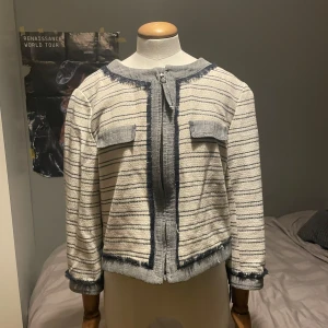 Randig croppad cardigan jacka med fransar - Säljer en ljusgrå och blå randig cardigan jacka med croppad passform och fransdetaljer längs kanterna. Jackan har trekvart ärmar och dekorativa fickor framtill. Perfekt för att lyfta en enkel outfit.