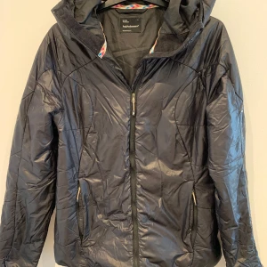 Svart pufferjacka från Peak Performance - Säljer en svart pufferjacka från Peak Performance med huva och dragkedja framtill. Jackan har två fickor med dragkedja och diskret logga på ärmen. Perfekt för kyliga dagar och enkel att matcha med olika outfits.