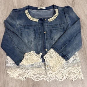 Jeansjacka med spets och pärldetaljer från Lexxy - Säljer en blå jeansjacka från Lexxy med dekorativa pärlor vid kragen och spetsdetaljer vid ärmslut och nederkant. Jackan har knappar framtill och två bröstfickor. Perfekt för dig som vill ha något unikt och sött i garderoben!