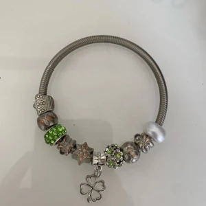 Silverfärgat armband med berlocker och fyrklöver - Säljer ett silverfärgat armband med flera berlocker i olika former och färger, bland annat gröna och vita detaljer samt en hängande fyrklöver. Perfekt accessoar för att ge din stil en personlig touch.