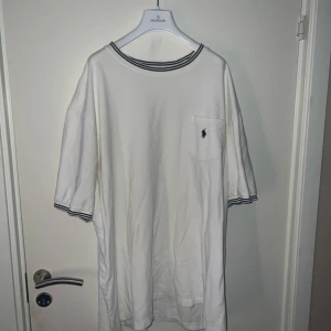 Vintage tröja eftertraktad  - Vit t-shirt från Polo Ralph Lauren med blå randiga detaljer vid krage och ärmslut. Klassisk passform med bröstficka och broderad logga. Perfekt för en clean och stilren look.