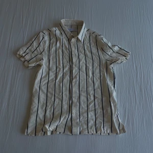 Vit och svart randig skjorta från Bershka - Kortärmad skjorta från Bershka i vit med svarta vertikala ränder. Klassisk krage och knäppning framtill. Perfekt för en avslappnad och stilren look.