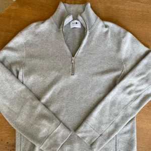 Ljusgrå halvzip-tröja från NN07 - Säljer en stilren ljusgrå halvzip-tröja från NN07. Tröjan har hög krage, dragkedja framtill och är tillverkad i mjukt material. Perfekt för lager-på-lager och enkel att matcha med olika outfits.