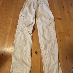 Vita cargopants med fickor - Vita cargopants med flera stora fickor på sidorna och röda detaljer på ficklocken. Byxorna har rak passform och är perfekta för en avslappnad stil. Materialet känns som bomull och de har en klassisk cargodesign.