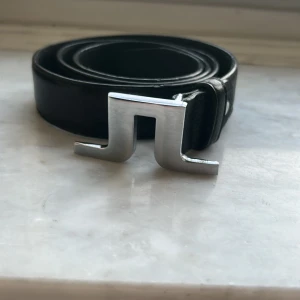 Svart läder bälte från J.Lindeberg - Säljer ett svart skärp från J.Lindeberg med ett stilrent, silverfärgat spänne i metall. Skärpet är gjort i skinn och har en modern design. Storlek 110/44. Perfekt accessoar för att lyfta din outfit. PRIS KAN DISKUTERAS 