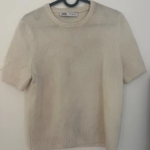 Beige stickad kortärmad tröja från Zara - Säljer en enkel och stilren beige stickad tröja från Zara. Tröjan har korta ärmar, rund halsringning och ribbade kanter vid ärmslut och nederlaget.