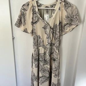 Somrig blommig klänning - Säljer en beige omlottklänning med svart blommigt mönster. Klänningen är midi längd. 