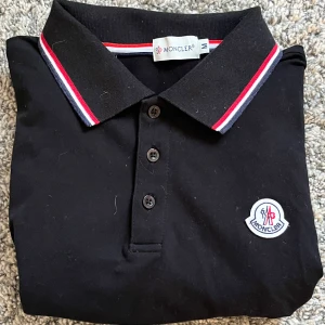 Moncler piké svart - Säljer en svart pikétröja från Moncler med klassisk krage och tre knappar framtill. Kragen och ärmsluten har röd, vit och blå randig detalj. Moncler-logga är broderad på bröstet. Använd bara två gånger. Vid frågor hör gärna av dig och få ett snabbt svar! 