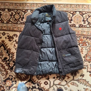 Svart dunväst med röd broderad detalj - Svart dunväst från Canada Goose med hög krage och dragkedja framtill. Västen har två stora fickor och en röd broderad logga på bröstet. Perfekt för lager-på-lager och håller dig varm under kyliga dagar.