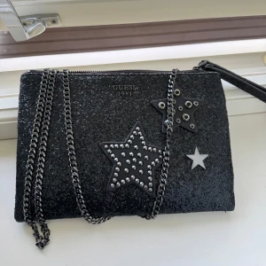 Svart glittrig axelväska från Guess - Säljer en svart glittrig axelväska från Guess med kedjerem och dekorativa stjärnor med nitar på framsidan. Väskan har dragkedja upptill och innerfoder med logotypmönster. Perfekt för att lyfta din outfit!