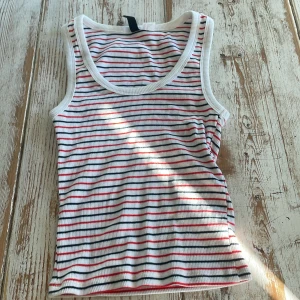 Randig ribbad linnetopp från H&M Divided - Säljer en vit ribbad linnetopp från H&M Divided med tunna horisontella ränder i svart och rött. Toppen har breda axelband och rundad halsringning. Perfekt för varma dagar eller som lager under en skjorta.