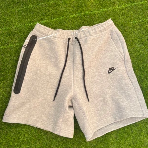 Grå shorts från Nike med svarta detaljer - Snygga grå shorts från Nike med svarta snören och dragkedja på ena fickan. Mjukt material och elastisk midja för skön passform. Perfekta för träning eller chill.